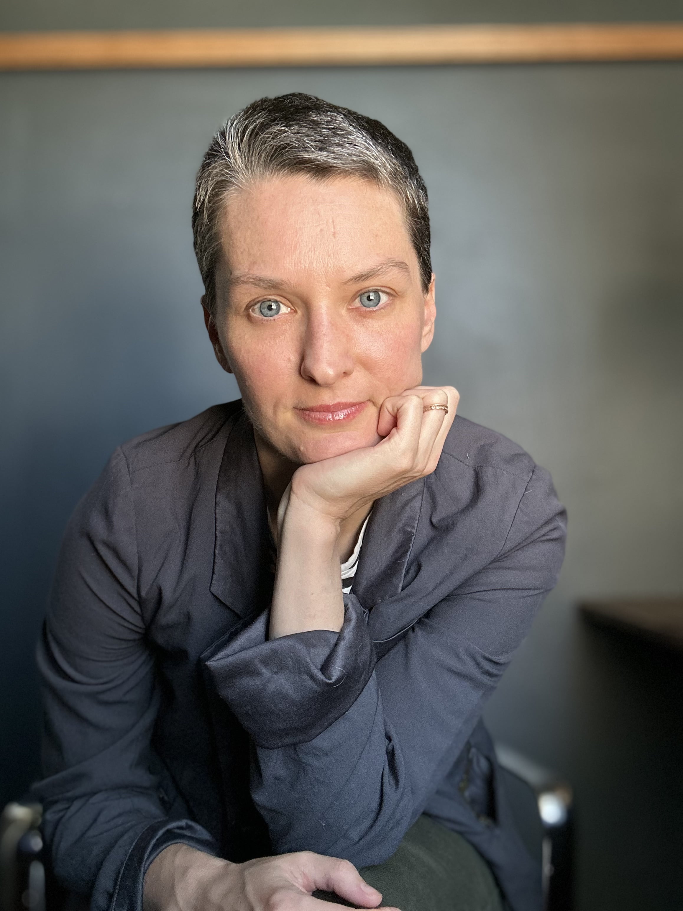 Person in grey shirt with short hair looking at camera and leaning chin on hand