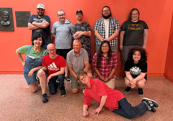 Group of adult theatre group posing in front of bright orange wall with director.