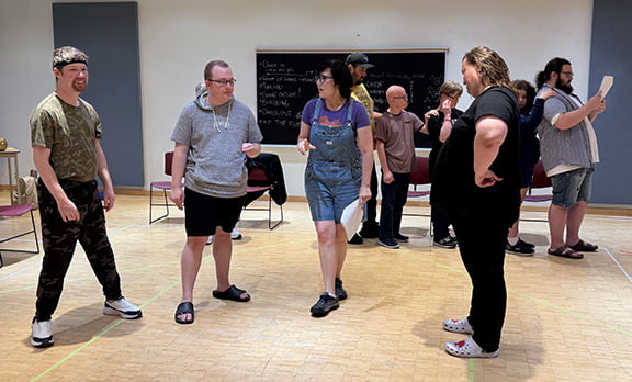 Group of adult students gathered around the director in a bright theatre rehearsal room with wood floors.
