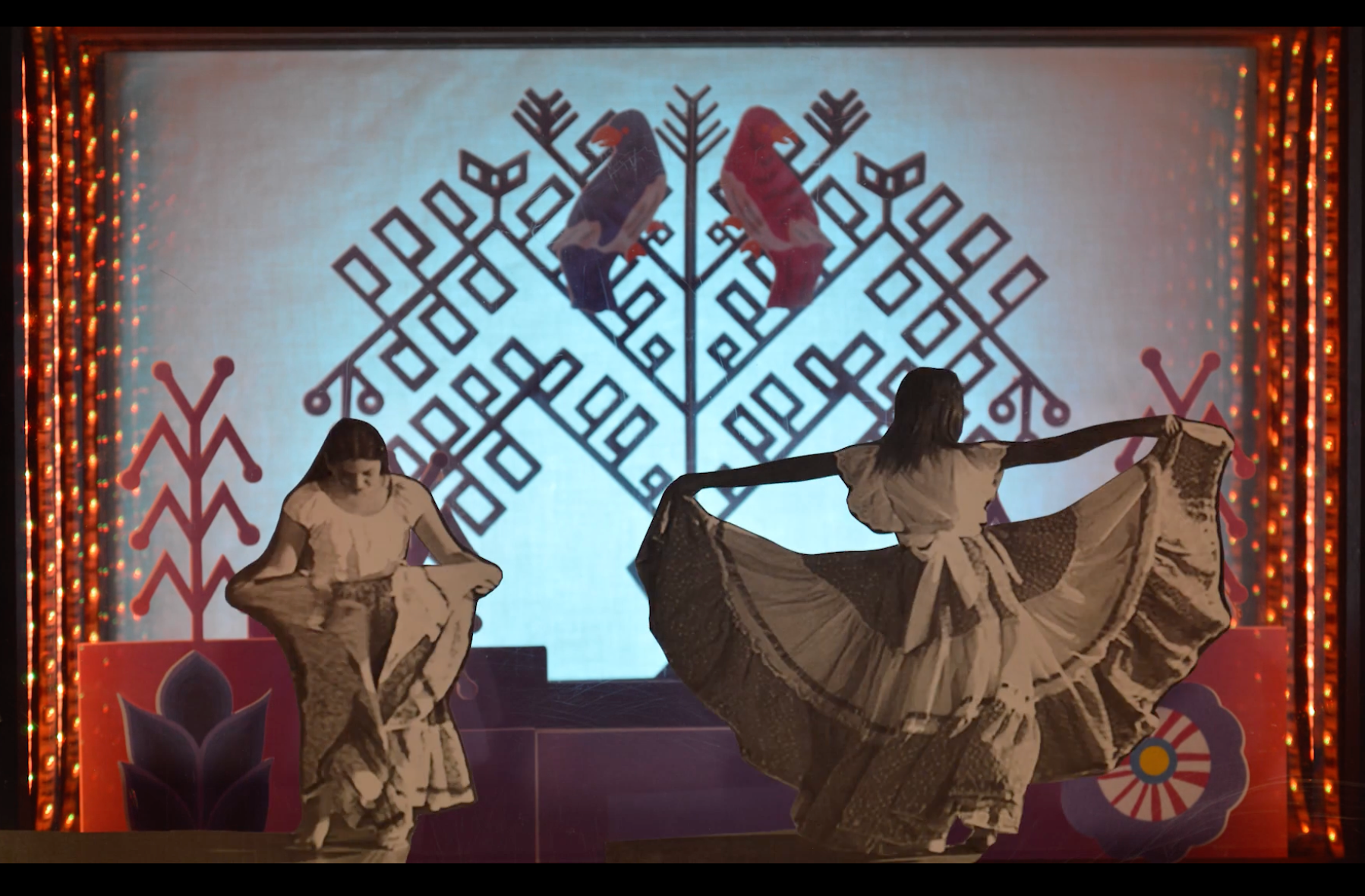 Two dancers in Folklorico skirts against paper-cut background.