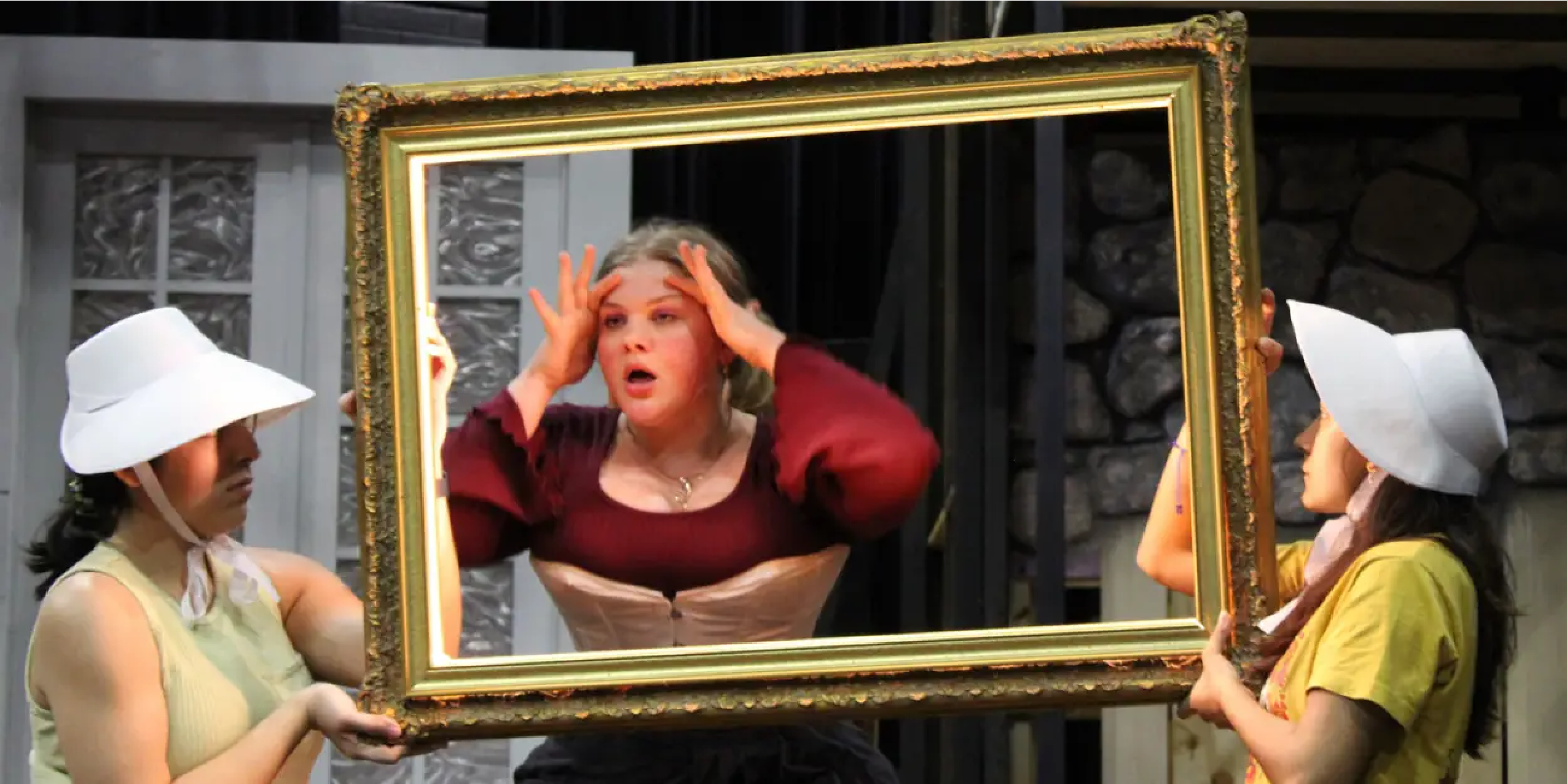 Three actors rehearse on stage, one looking through a frame as if it were a mirror, two in white bonnets