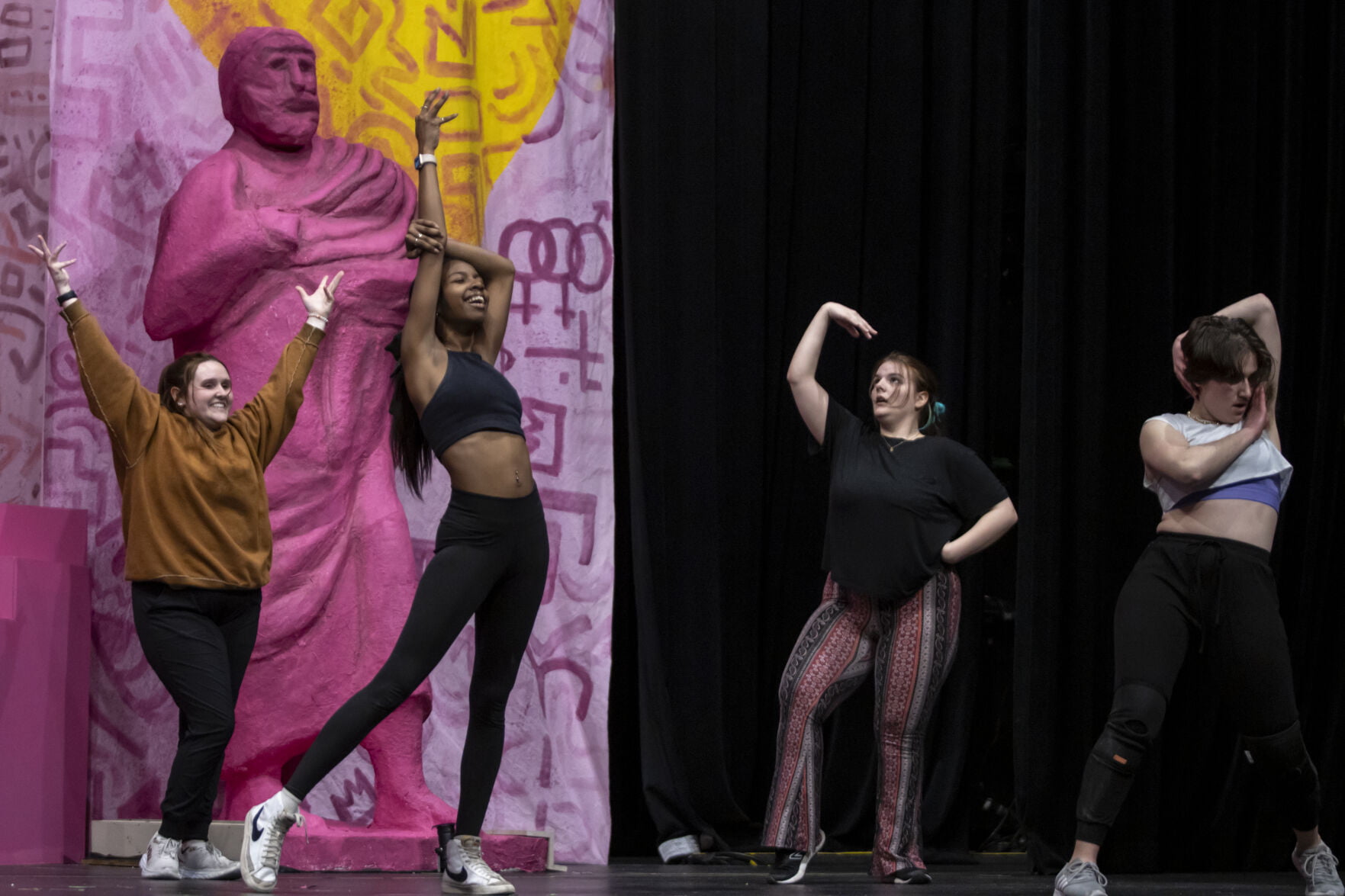 Four performers striking a pose in front of massive pink greek statue.