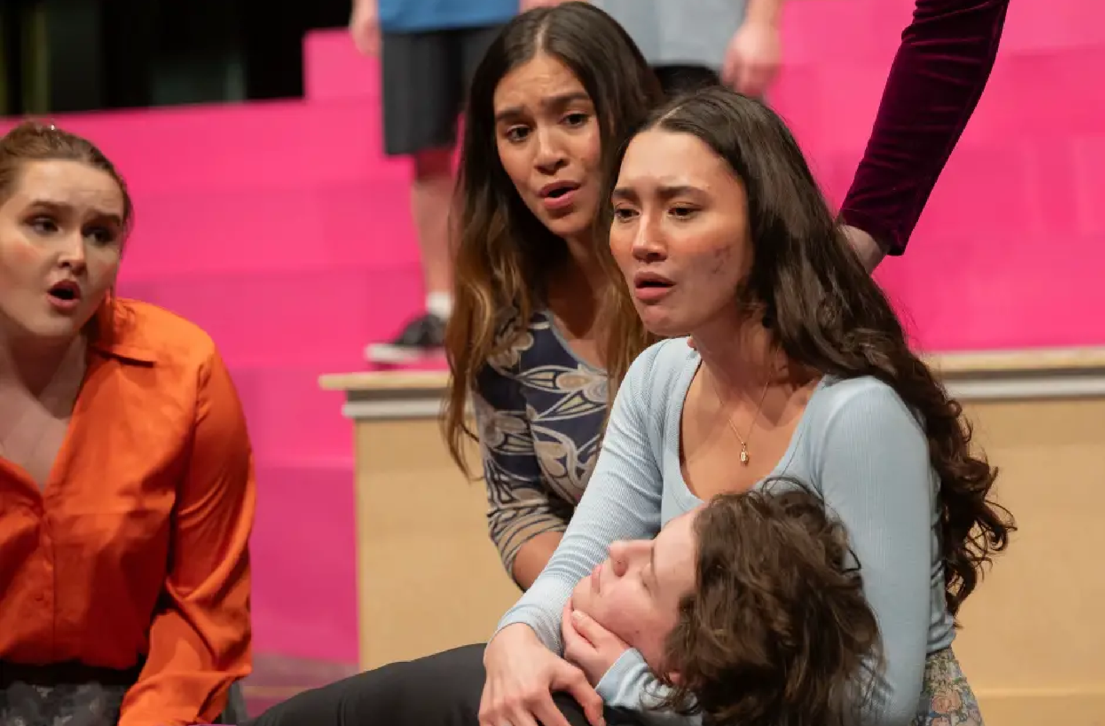 Three women characters singing over a dying male character, rehearsing on a stage with bright pink background.