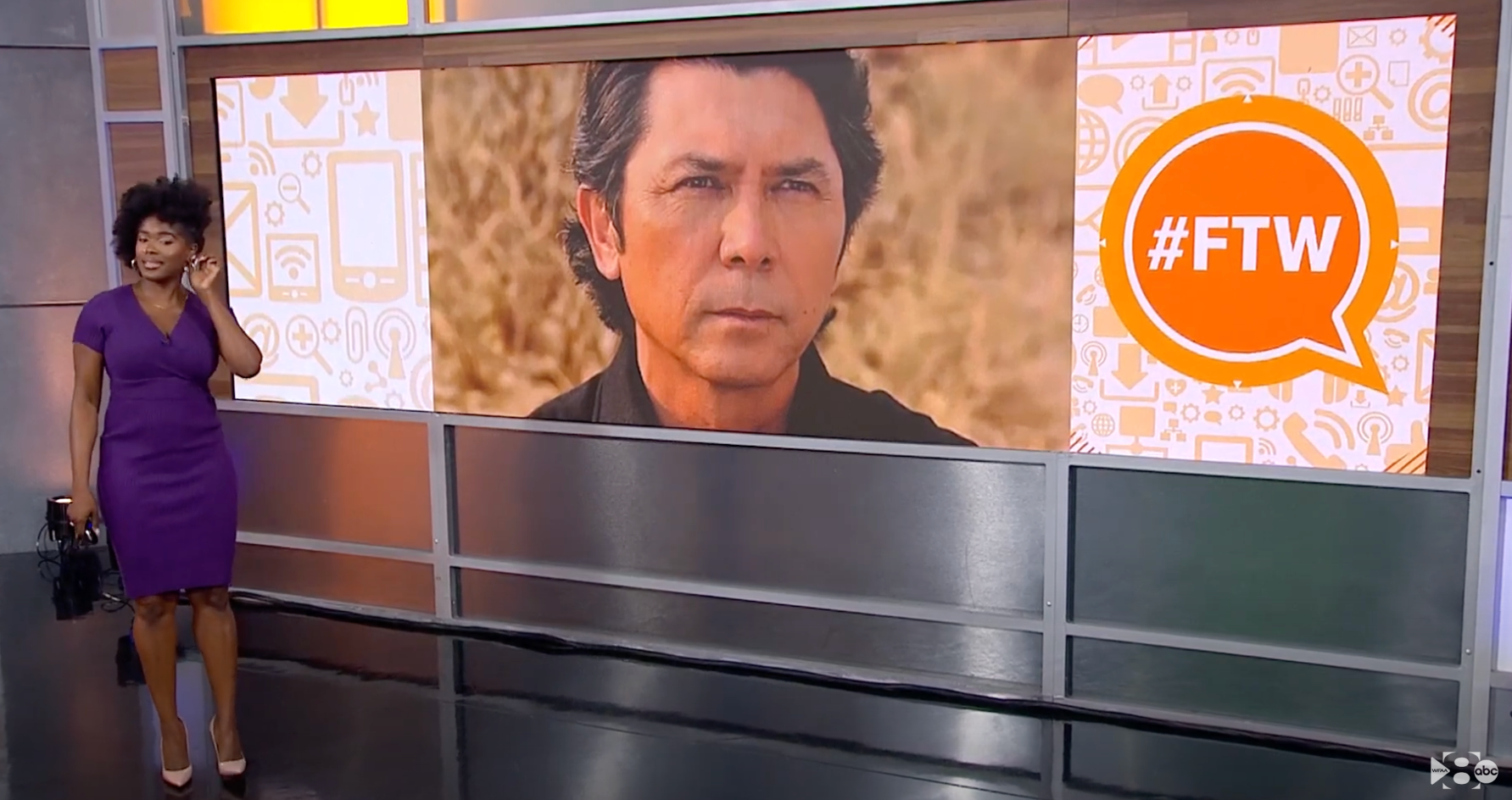 Newscaster in purple dress next to screen image of actor Lou Diamond Phillips