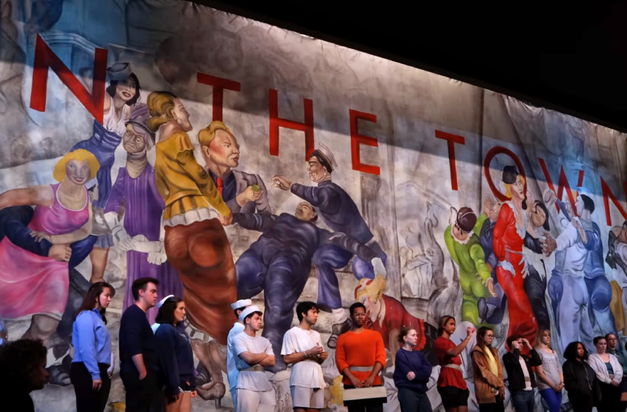 Actors lined up in front of elaborately painted backdrop of people from a Paul Cadmus painting