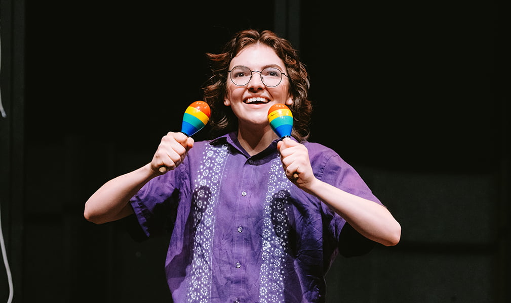 Smiling performer in purple shirt with embroidered look holding colorful maracas