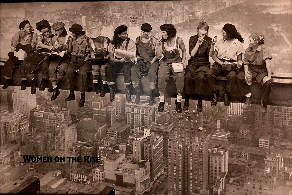Sepia color of several women dressed as 1940-1950s construction workers sitting on steel beam with a cityscape below and behind them. The title Women On the Rise is in the bottom left corner." _languageinserted="true