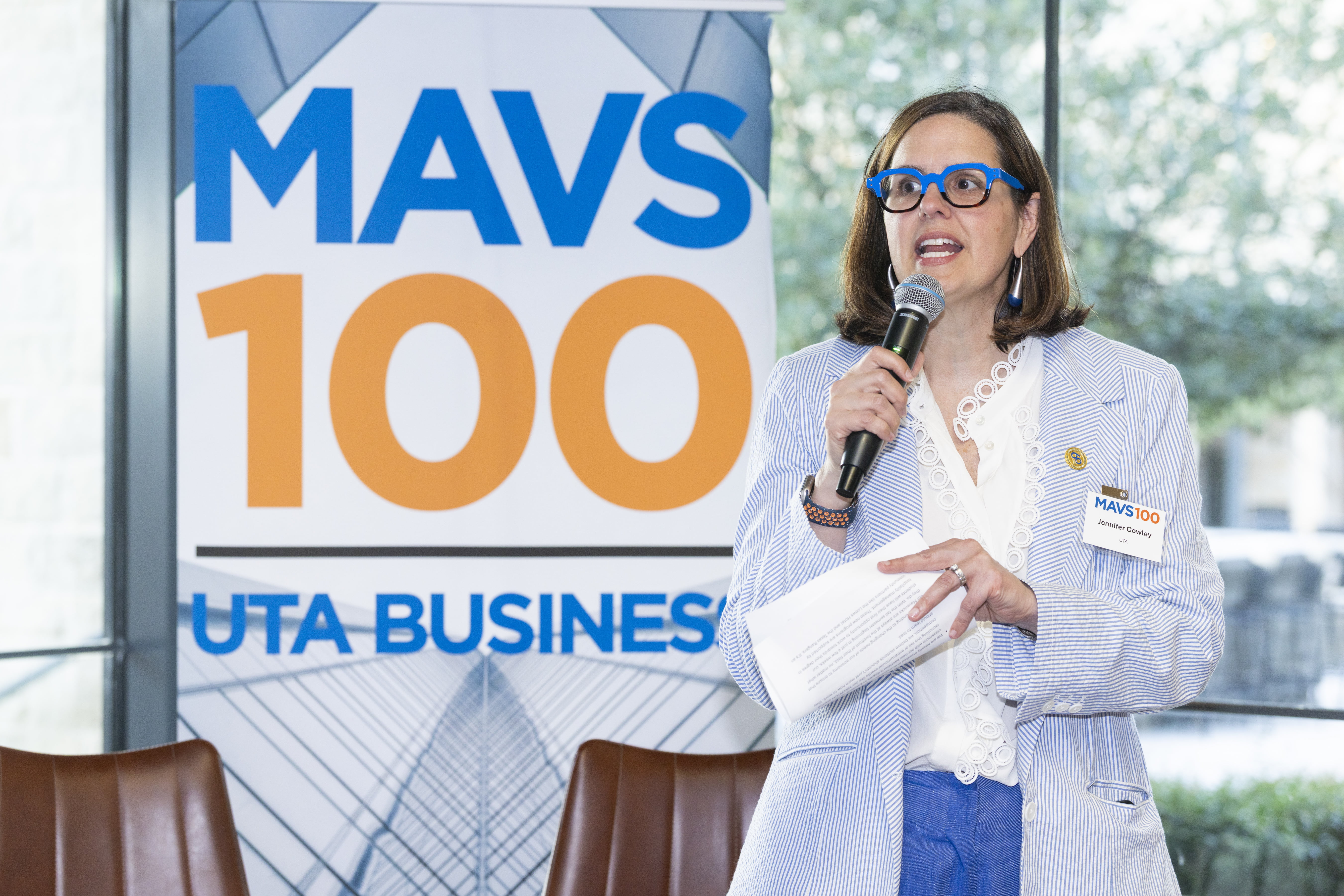 UTA President Jennifer Cowley speaks into a microphone. Behind her is a banner advertising MAVS100. 