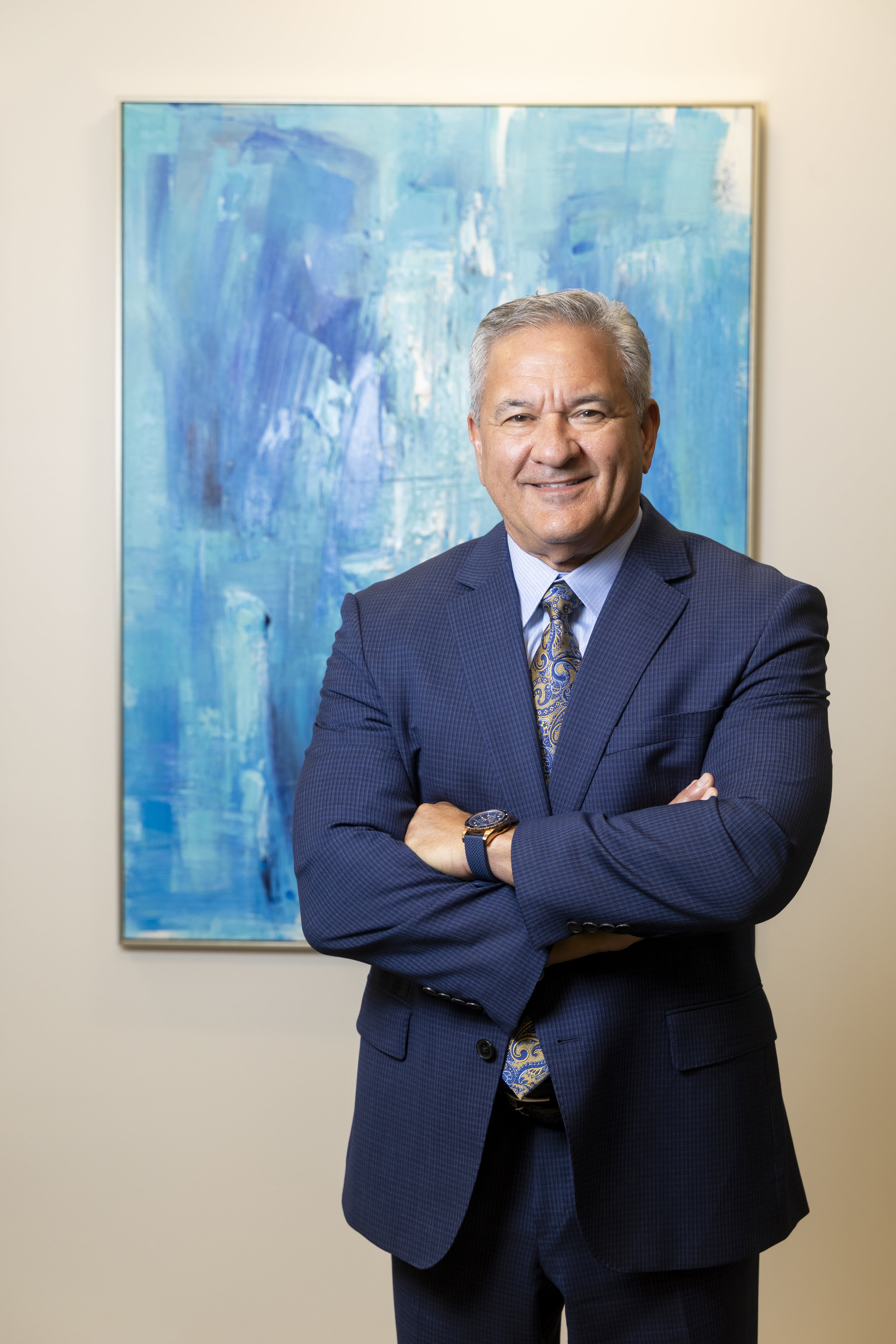 Greg Barron stands in front of a blue abstract painting, smiling while crossing his arms. 