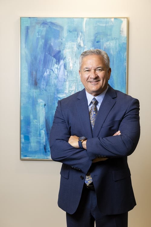 Greg Barron stands in front of a blue abstract painting, smiling while crossing his arms.
