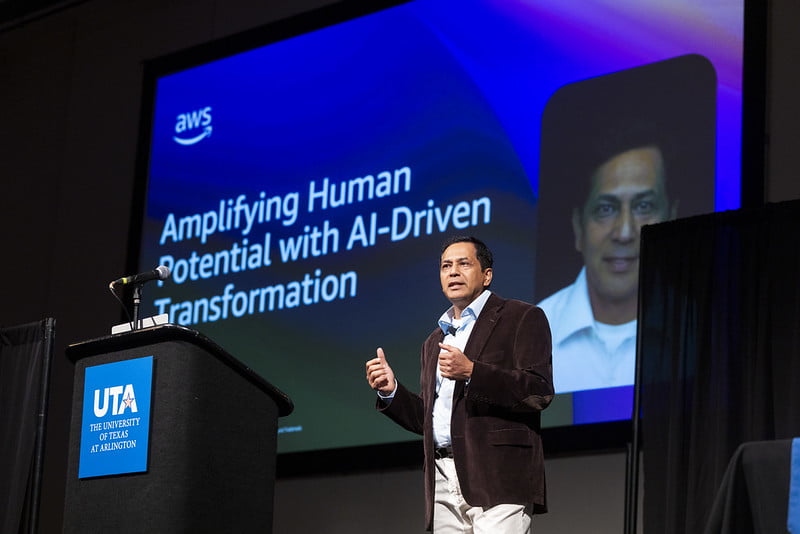 Heddese speaks to attendees from a podium. Behind him, his powerpoint features his headshot and the words "Amplifying Human Potential with AI-Driven Transformation"