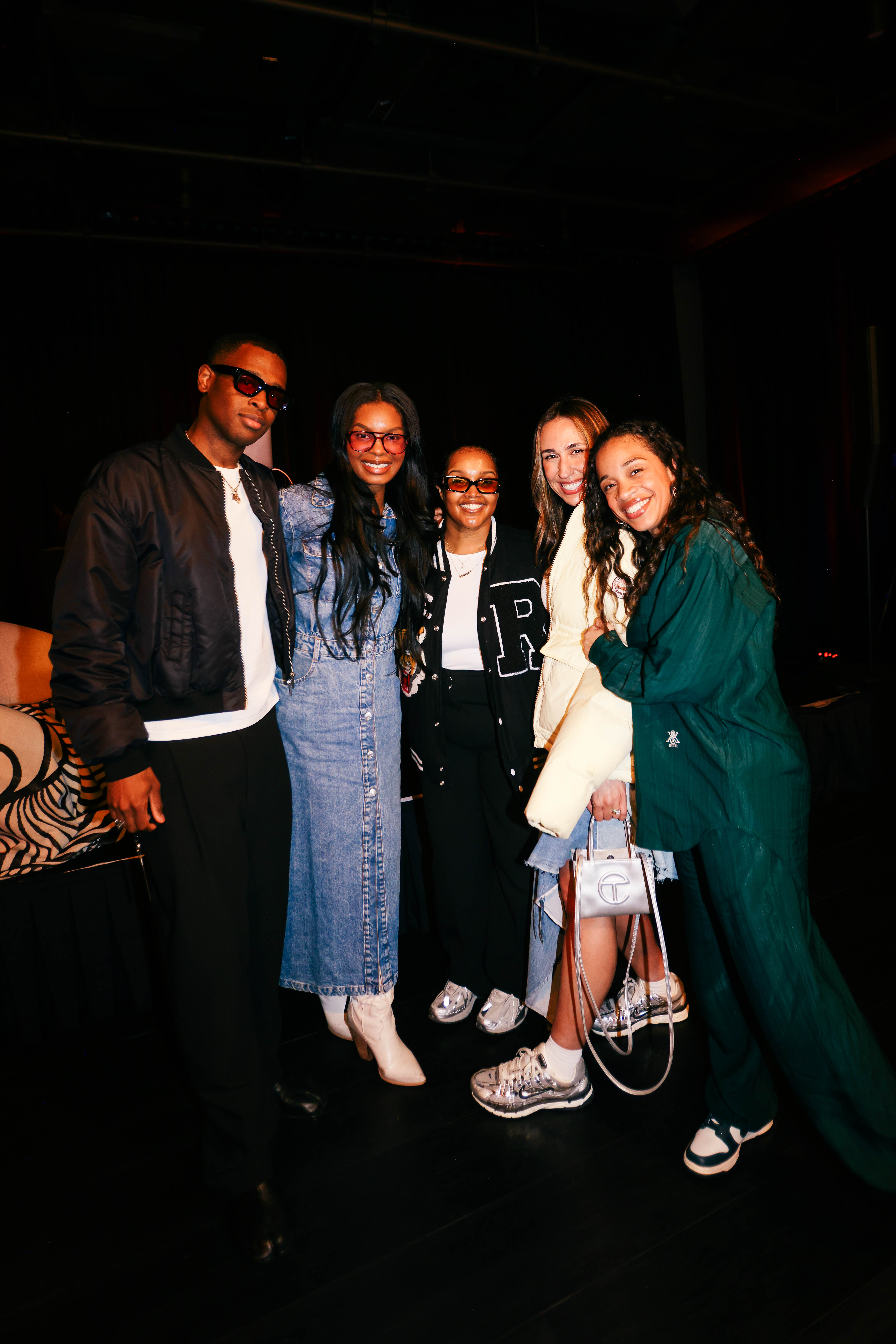 Five people stand close together smiling for a group photo in a dimly lit event space. From left to right: a man in sunglasses and a black bomber jacket; a woman in a denim maxi dress and white boots; a woman in a black and white varsity jacket; a woman in a light jacket and denim skirt holding a silver Telfar bag; and a woman in a green tracksuit and Nike sneakers.