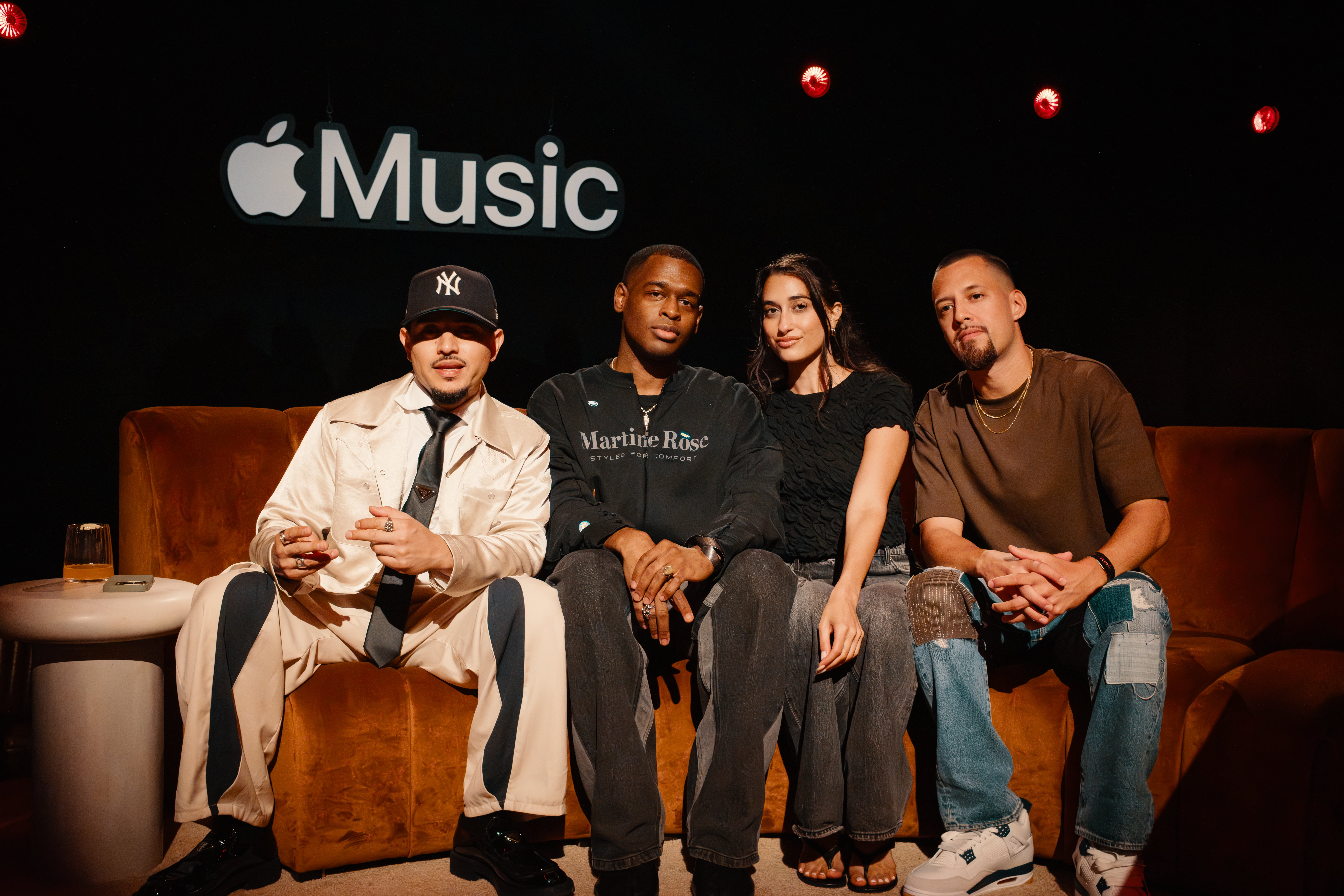 Four individuals sit on a burnt orange velvet couch in front of an "Apple Music" sign on a black backdrop. From left to right: a man in a cream outfit and black Yankees cap flashes a peace sign; a man in a black "Martine Rose" sweatshirt sits with his hands clasped; a woman in a black textured top and dark jeans smiles slightly; and a man in a brown shirt and patchwork jeans rests his hands in his lap.