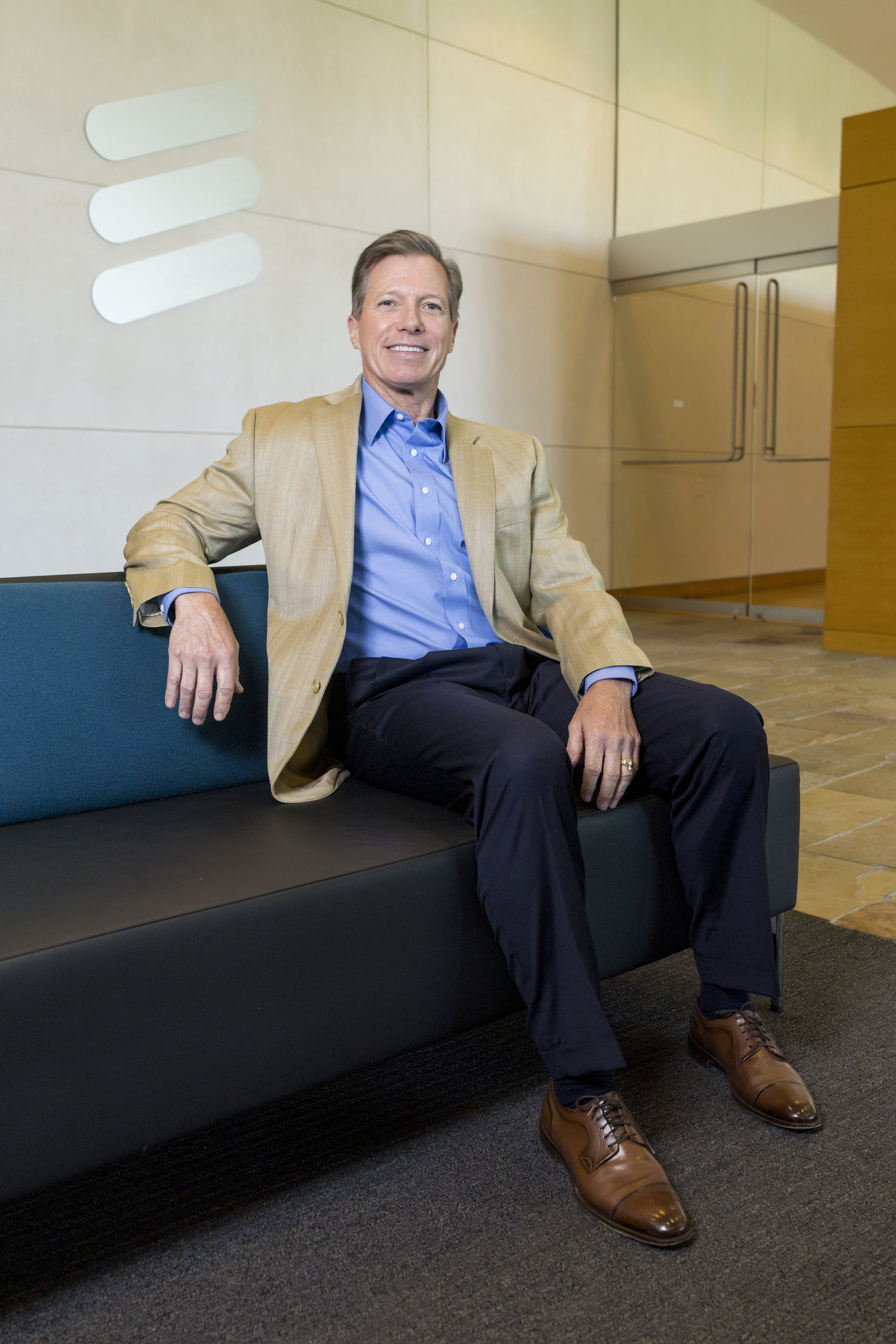 Brian White poses for a headshot. He is seated, leaning against the back of a blue couch. Behind him, the Ericsson logo is visible. Photo by Sharon Ellman. 