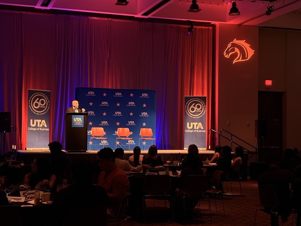 A speaker stands at a podium with the UTA College of Business logo, addressing an audience seated at tables. The stage backdrop features banners with “60 Years” and UTA branding, illuminated with orange and blue lights.