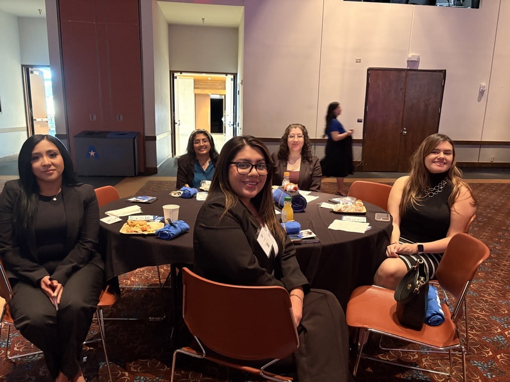 A group of five women dressed in business attire sit around a round table at a professional event in the University of Texas at Arlington’s College of Business. Plates of food, name tags, and event materials are visible on the table. The room has brown flooring and neutral walls with open doors in the background.