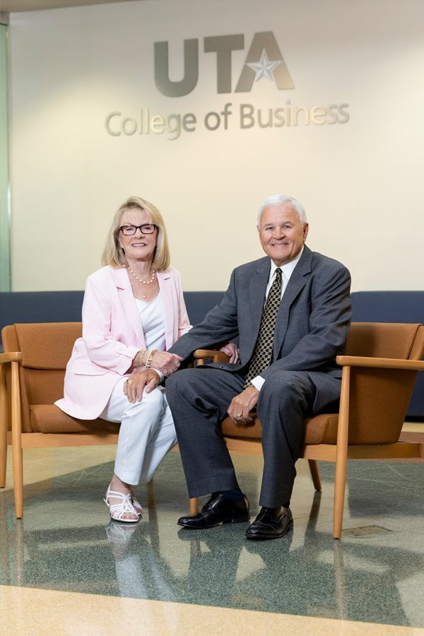 Jimmy and Cathy pose for a photo. Cathy sits on the left, wearing a pink jacket and white pants, holding Jimmy on the right who wears a dark suit.