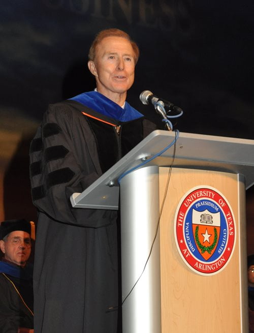 John Merrill stands at a podium featuring the UTA seal on it. He is speaking into the microphone while wearing doctoral robes.