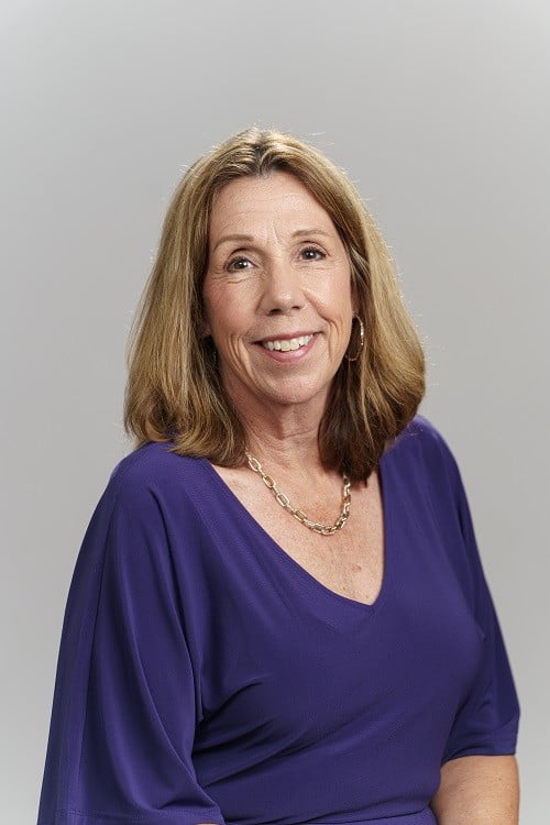 Kelly Roberts smiles for a headshot, wearing a purple dress against a grey background. 