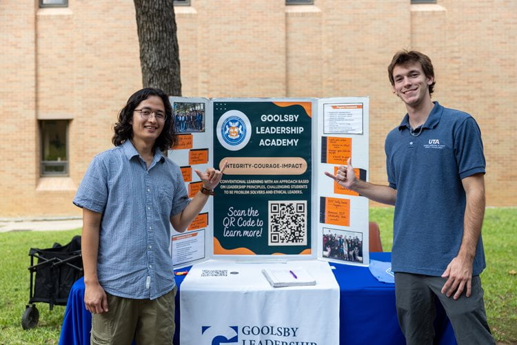 Two students give a Mav Up hand sign in front of a board advertising the Goolsby Leadership Academy at the UTA College of Business Student Organization Fair on September 11, 2024. (Photo by Jalen Larry)