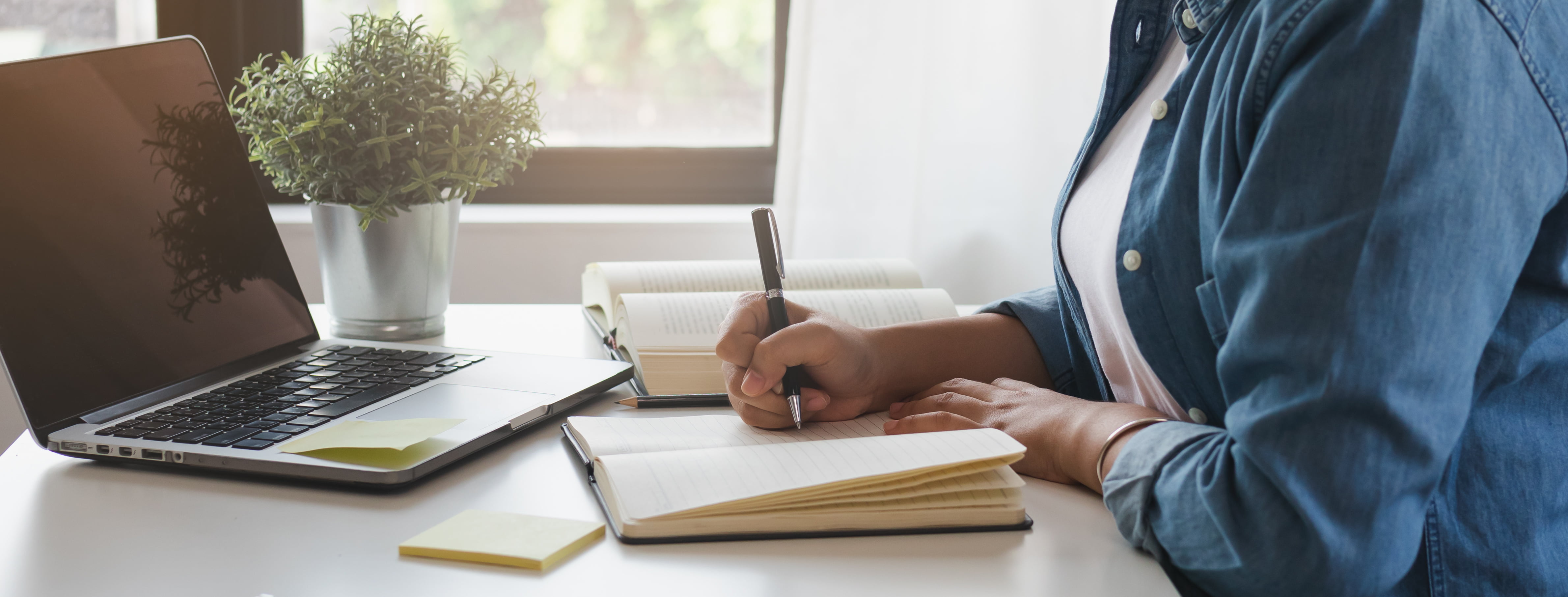 An unidentified person writes in a journal in front of an open laptop.