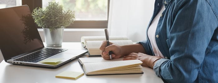 An unidentified person writes in a journal in front of an open laptop.