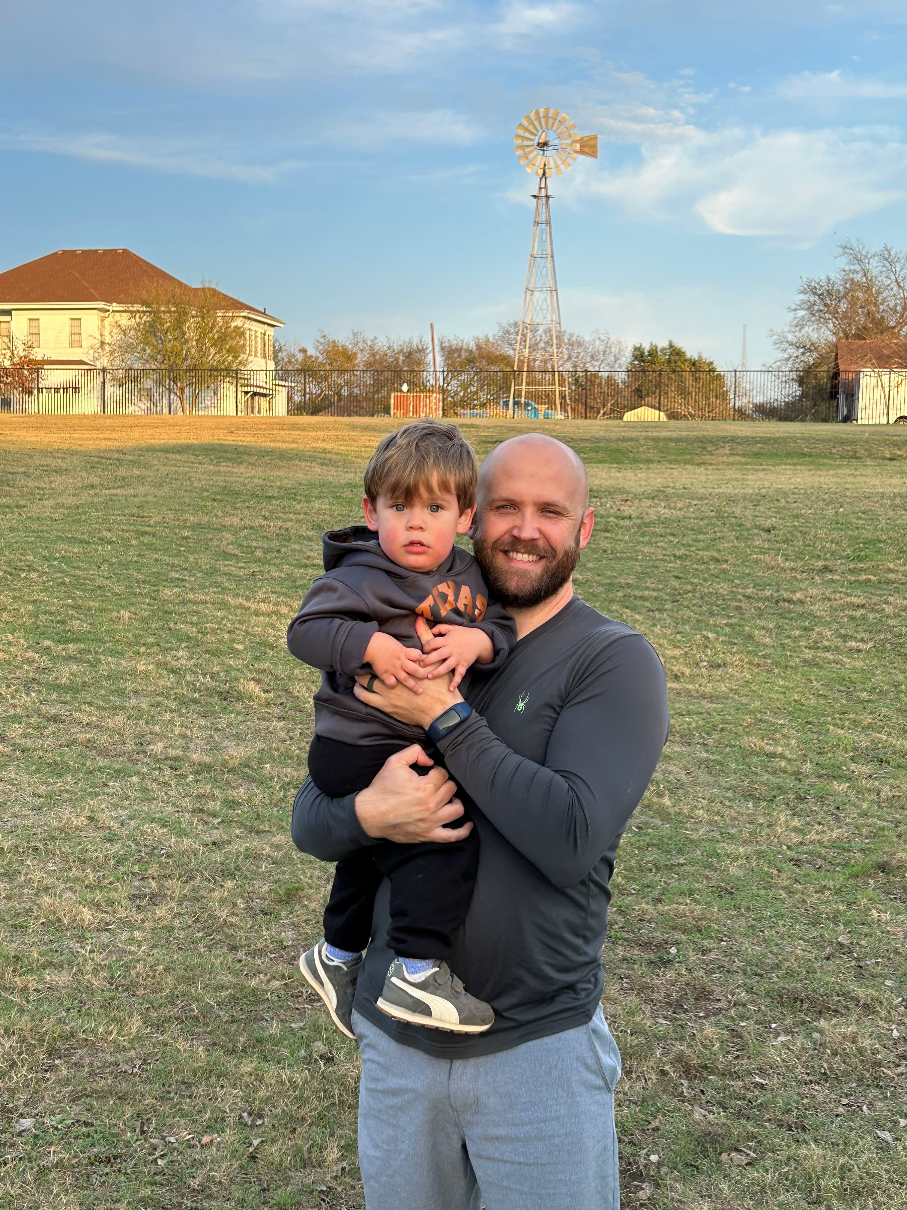 Scott Elbert poses for a picture with his toddler, Henry. Outside on a green field. 