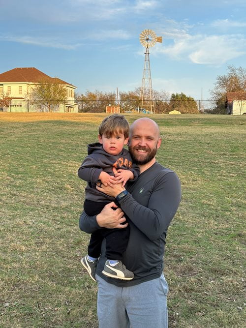 Scott Elbert poses for a picture with his toddler, Henry. Outside on a green field.