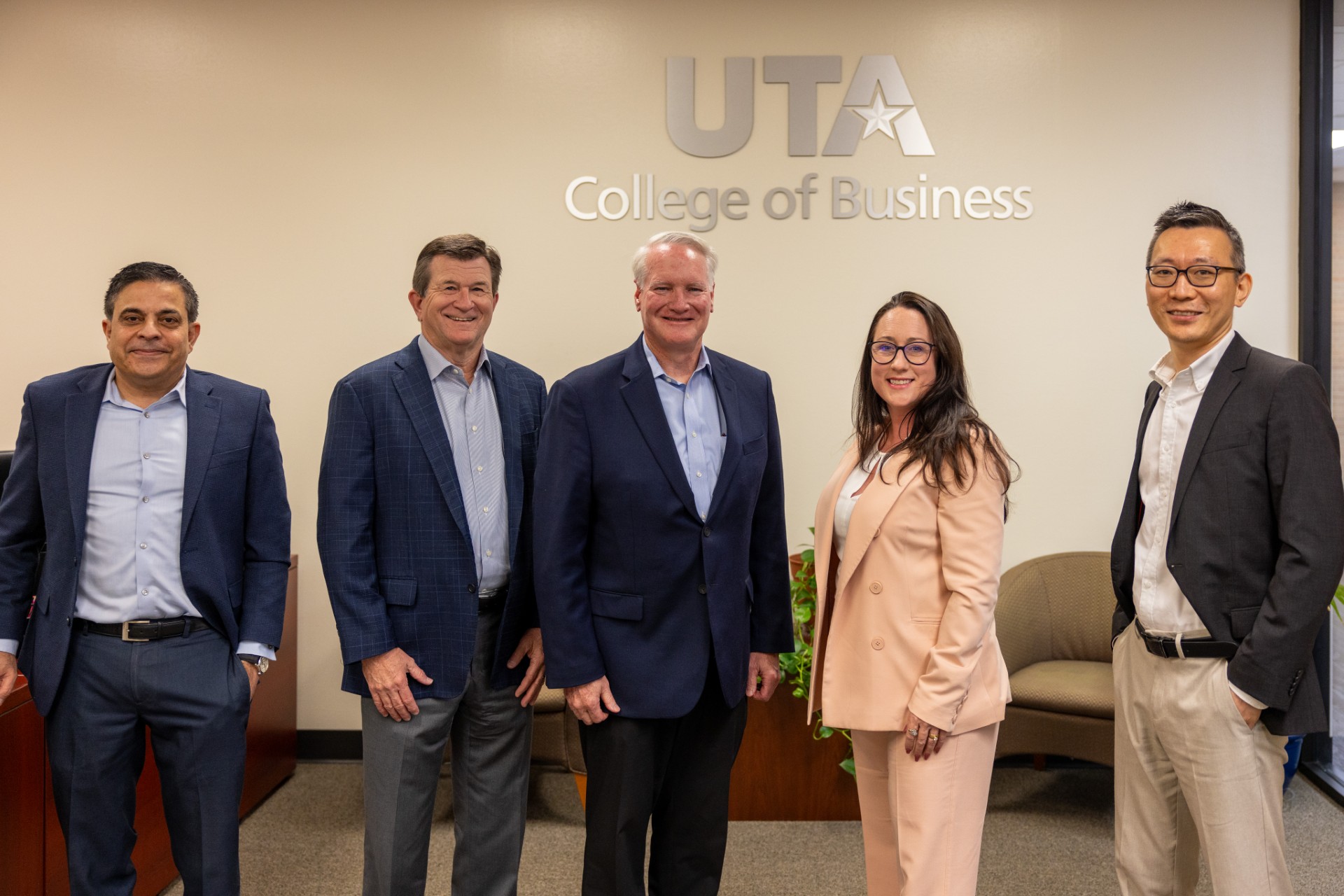 From left to right: Dr. Sanjiv Sabherwal, Dean Harry Dombroski, Rick Jenkins, Lacy Samples and Dr. Ten Wang. The group poses together for a photo, with the UTA College of Business logo on the wall behind them. 