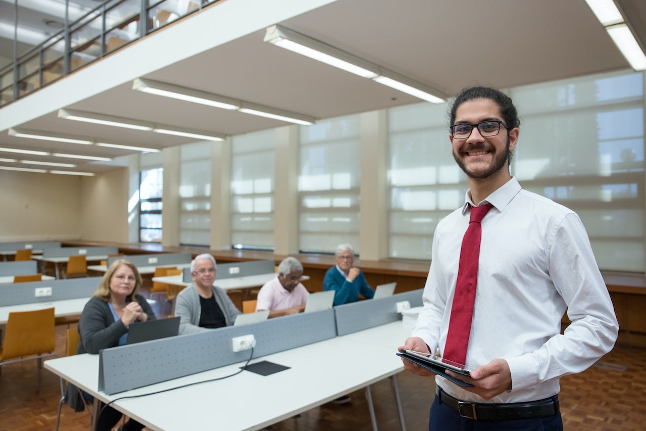 Teacher holds a tablet in a library classroom of senior citizens working on laptops.