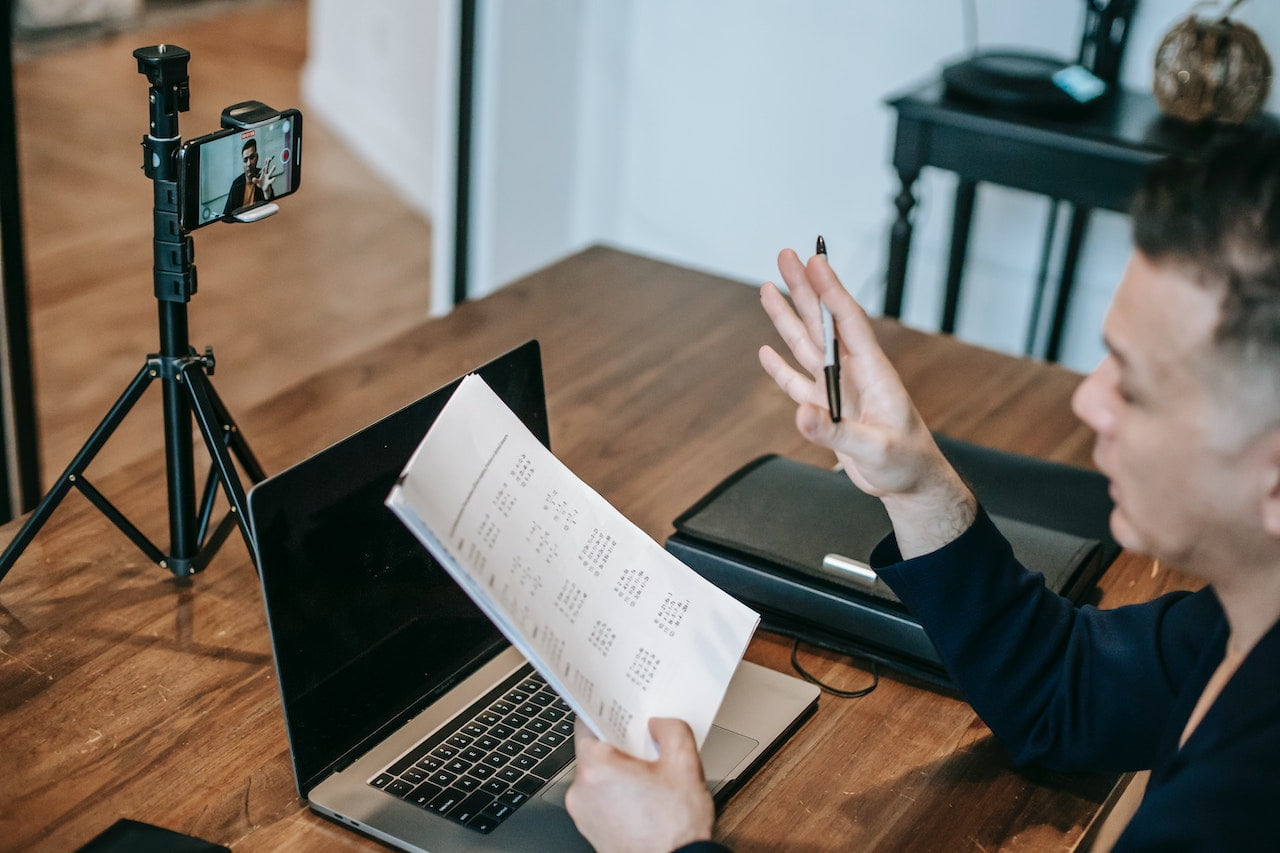 A teacher works on a laptop and a records a video with a smartphone