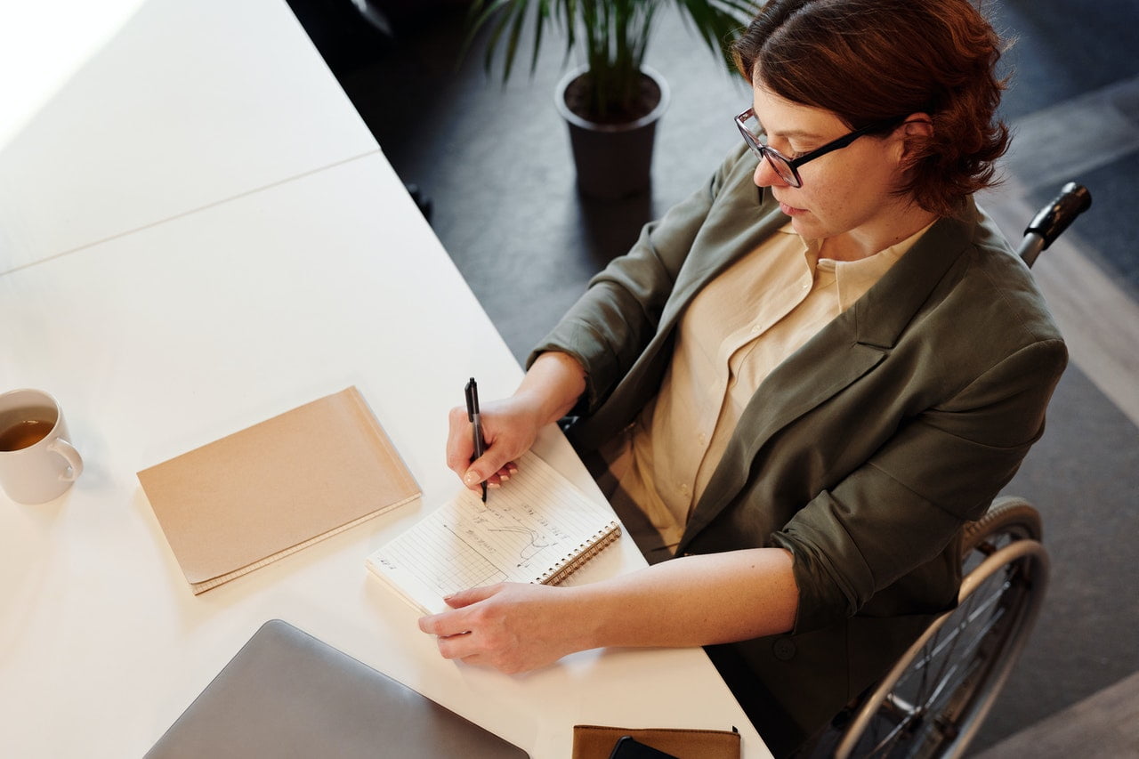 A woman in a wheelchair sits at a desk and writes on a piece of paper