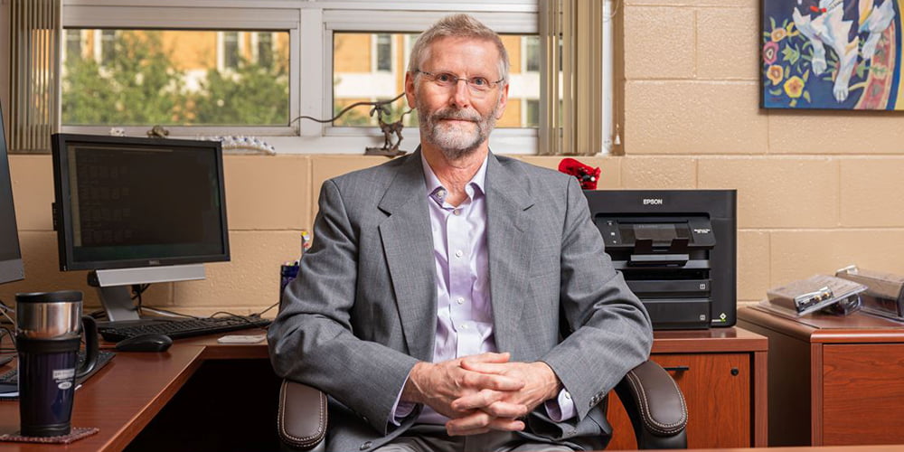 A man with white hair sits smiling at his desk." _languageinserted="true