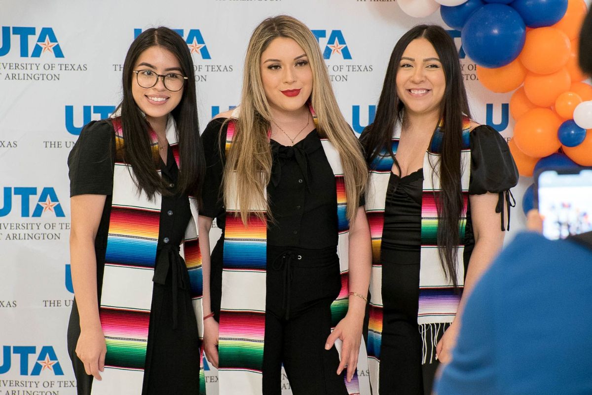 Three UTA students wearing sarape stoles, posing in front of UTA background