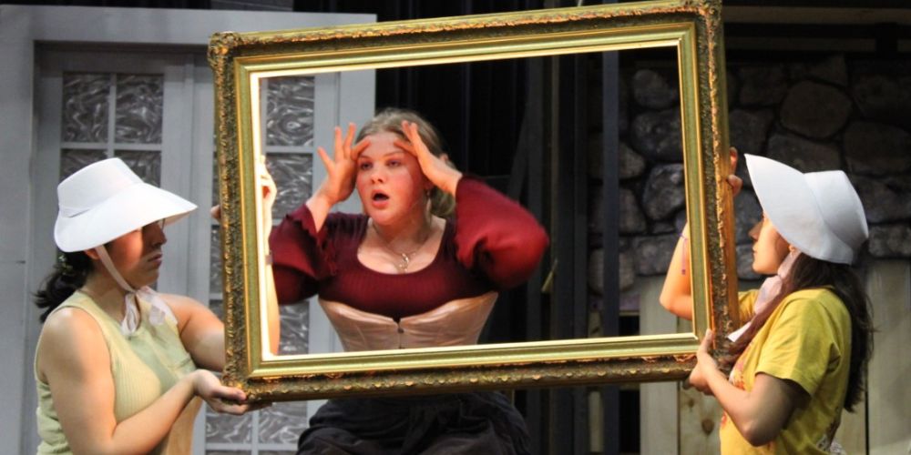 Two women in profile holding an empty picture frame while another woman looks through it 