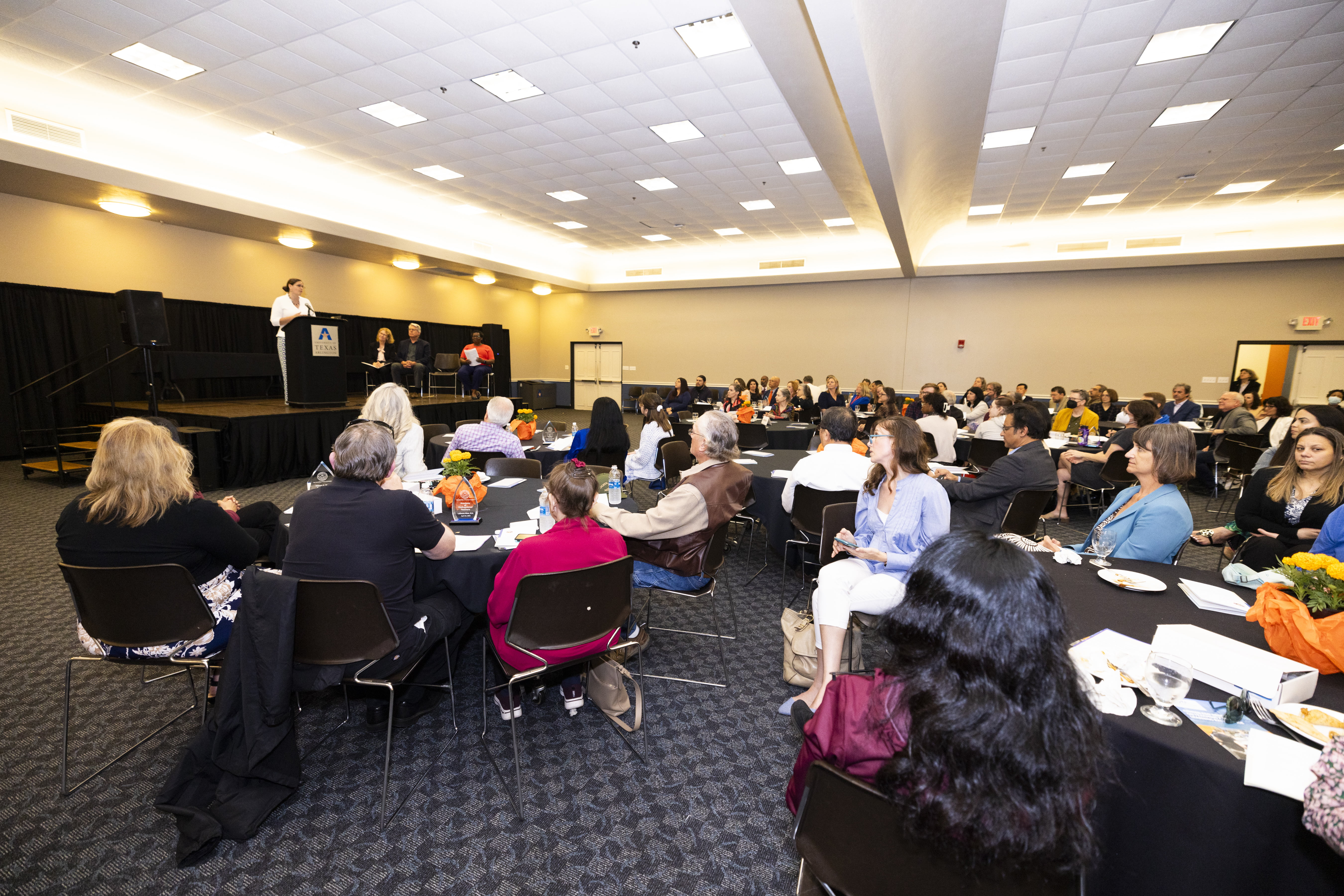 A group of people in a room, sitting at tables looking a speaker on a podium. 