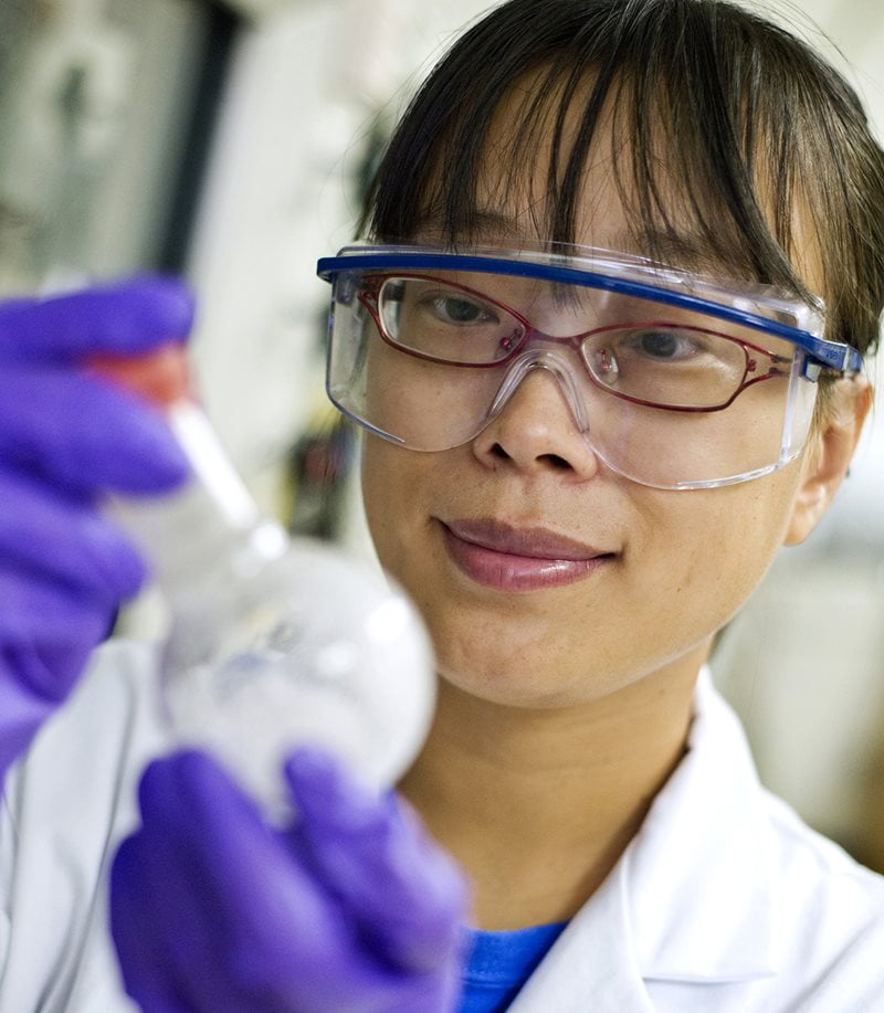 A graduate student wearing safety goggles and gloves holding a test tube to observe a chemical reaction in a laboratory in the Chemistry & Physics Building.