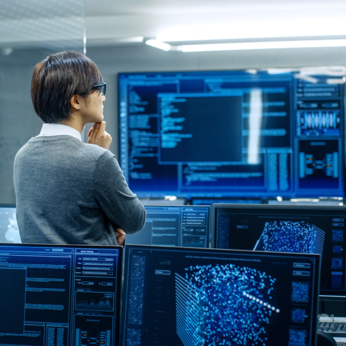 A researcher in a glasses-filled data center analyzing complex blue digital information displays.