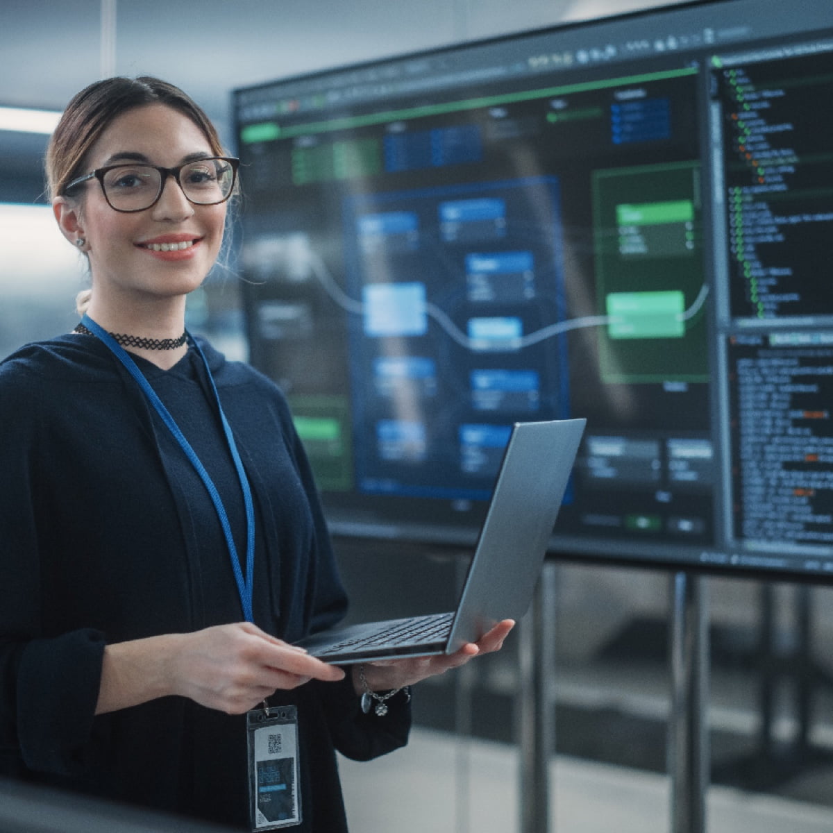 A smiling female IT professional holding a laptop in front of a digital dashboard.