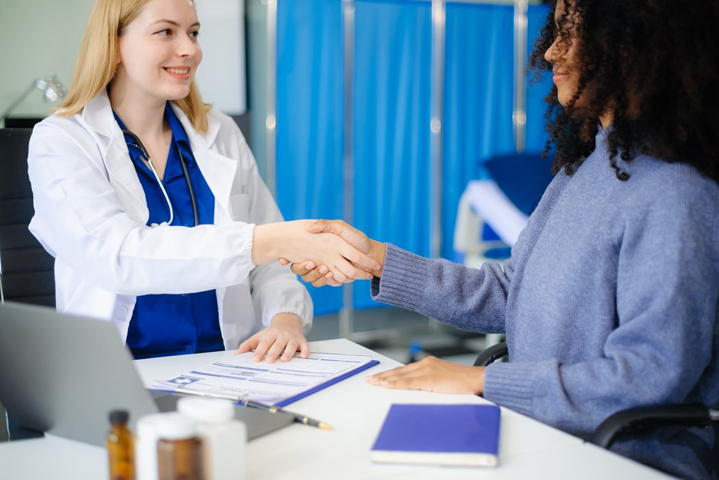 A doctor in a white coat and a patient in a sweater shake hands across a desk, creating a warm, professional atmosphere. A clipboard and laptop are visible.