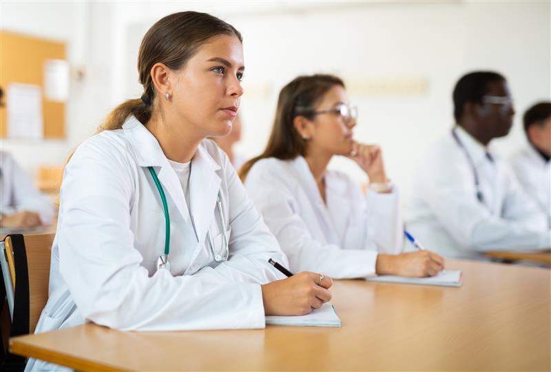 Medical students in white lab coats attentively taking notes in a classroom setting, with desks and educational materials visible.