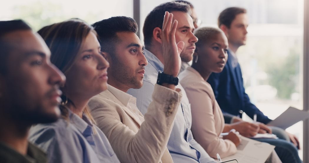 A diverse group of people sitting in a row, attentively engaged. One person raises a hand, suggesting they have a question or comment. The setting appears formal.