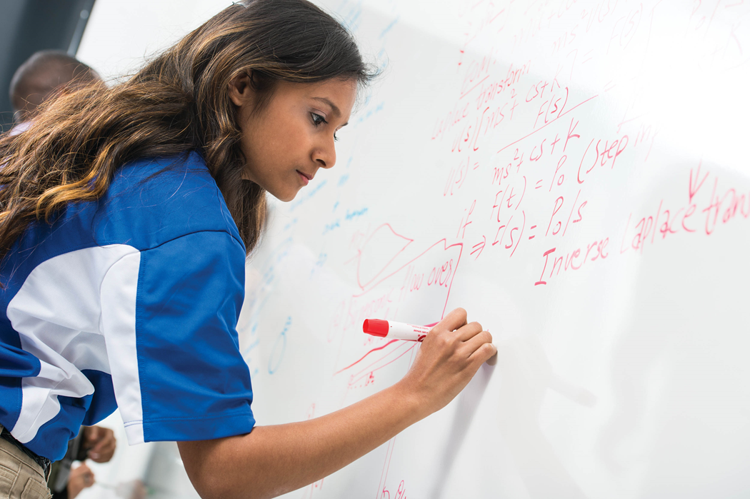A female math student uses a marker to write an equation on a whiteboard.