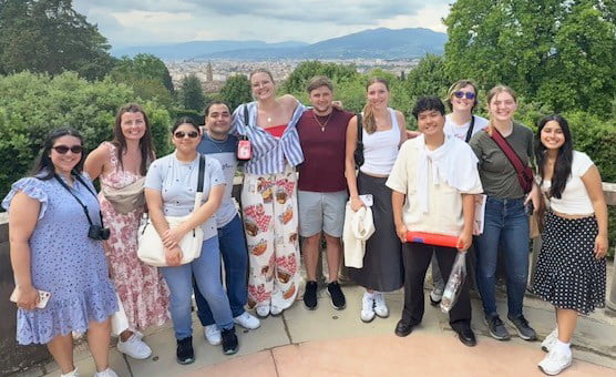 A diverse group of 12 people poses together outdoors with a scenic backdrop of mountains and greenery.