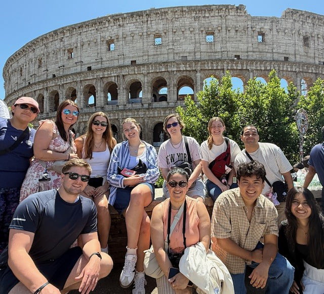 A group of people poses in front of the Colosseum in Rome, surrounded by greenery, enjoying a sunny day.