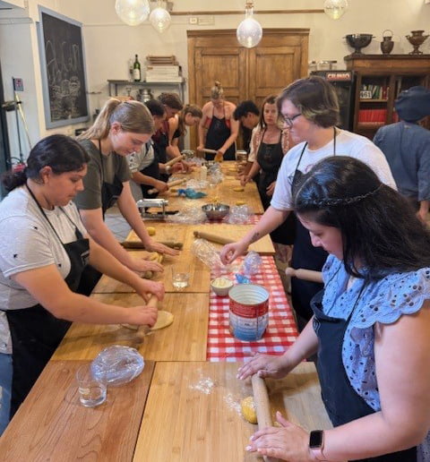 A group of people in aprons work at communal tables, preparing dough and ingredients in a cozy cooking class setting.