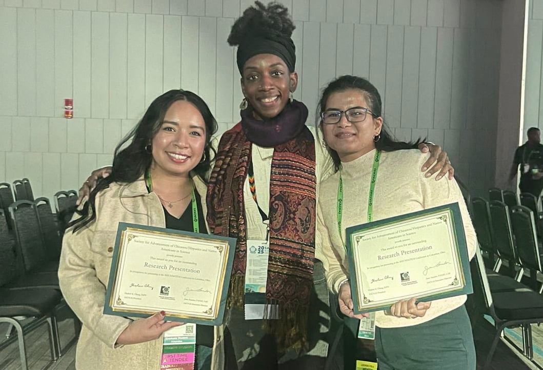 Three smiling women pose together, celebrating a research achievement. Angelica Torres (left) and Trilotma Sen (right) are holding certificates recognizing their outstanding research presentations in cell/molecular biology and life sciences.