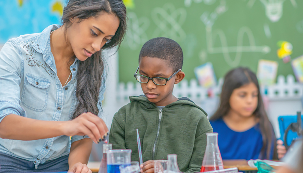 An elementary school teacher and a young student observing a chemical reaction in a classroom.