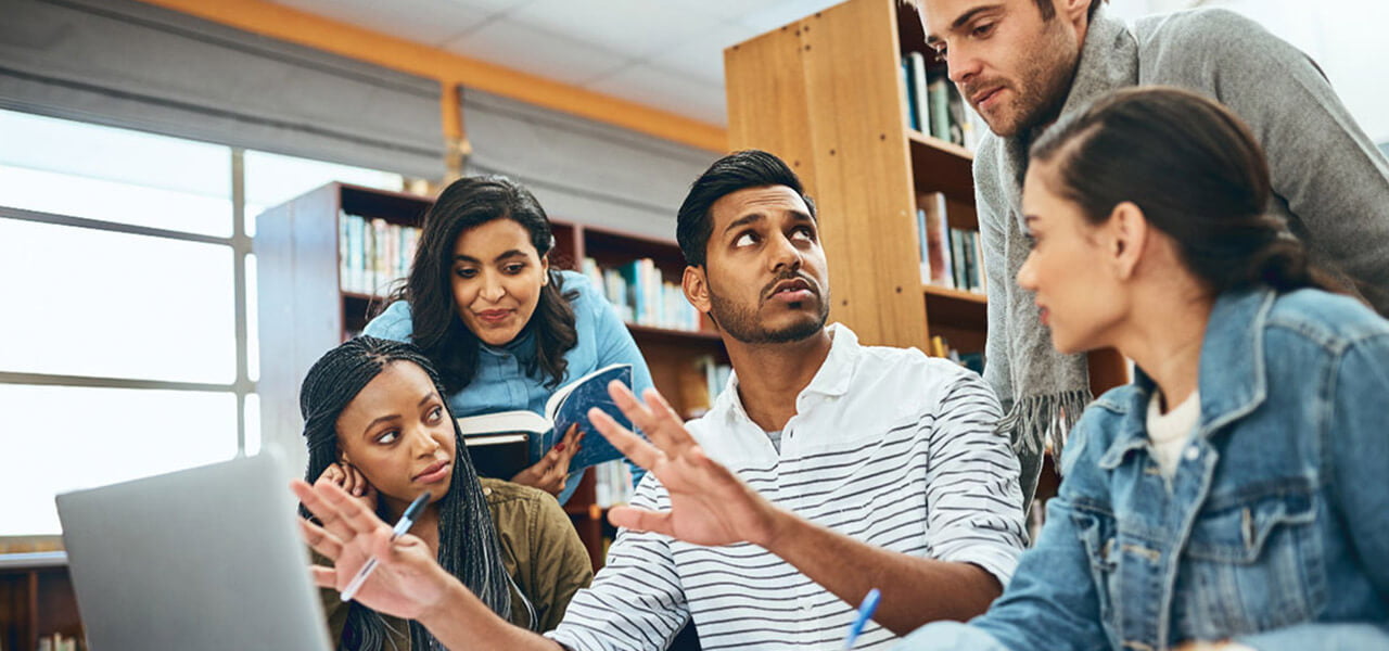 group of 5 students talking in front of a laptop in the library