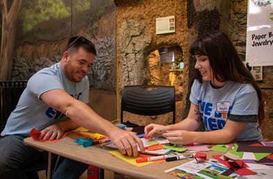 Two volunteers making jewelry with colored paper and markers
